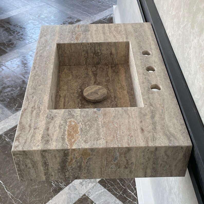 May include: A rectangular, stone bathroom sink with a built-in basin and a circular drain cover. The sink has three faucet holes and a natural, mottled color scheme of beige, brown, and gray.