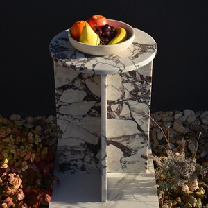 May include: A marble side table with a round top and a single support. A bowl of fruit, including an orange, apple, pear, grapes, and banana, sits on the table. The table has a white and purple veined pattern.