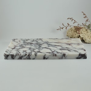 May include: A rectangular marble cutting board with a white base and dark purple veining. The board is smooth and polished, with a natural stone pattern. The board is suitable for kitchen use.