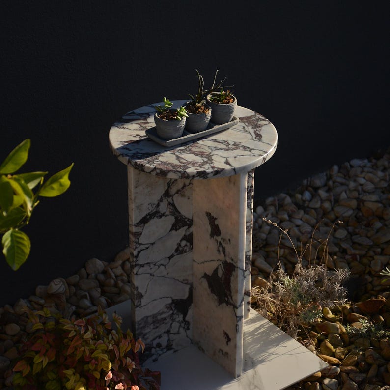 May include: A marble-topped pedestal table with a round surface and a rectangular base. The table features a white and purple veined pattern. Three small potted plants sit on a gray tray on top of the table. The table is outdoors.
