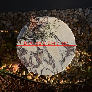 May include: A round marble table with a diameter of 16 inches. The table features a white and burgundy veined pattern. Dried flowers are placed on the table's surface. The table is set on a bed of small stones.
