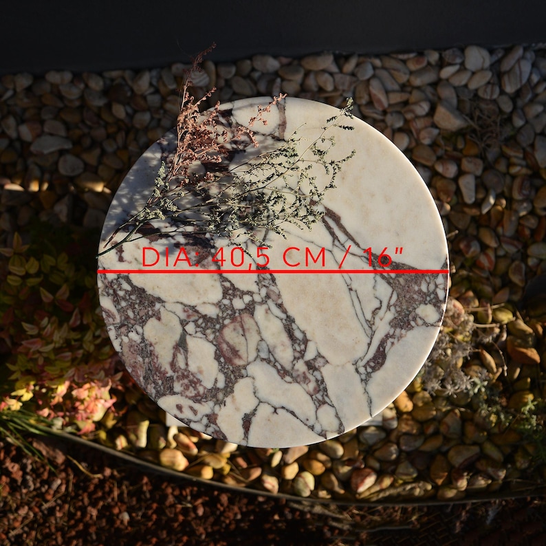 May include: A round marble table with a diameter of 16 inches. The table features a white and burgundy veined pattern. Dried flowers are placed on the table's surface. The table is set on a bed of small stones.