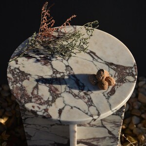 May include: A small, round marble table with a white and burgundy patterned top. The table has a single, slender leg. Several walnuts and dried sprigs of greenery rest on the tabletop. The background is dark.