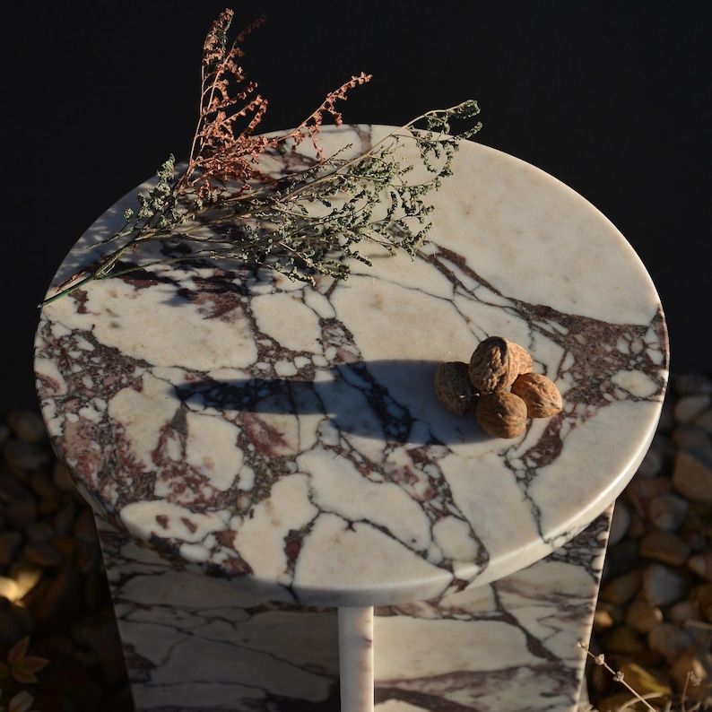 May include: A small, round marble table with a white and burgundy patterned top. The table has a single, slender leg. Several walnuts and dried sprigs of greenery rest on the tabletop. The background is dark.