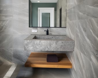 May include: A modern bathroom with gray stone walls and flooring. A rectangular gray stone sink is mounted on the wall above a wooden shelf. A black faucet and mirror are above the sink. A dark purple towel rests on the shelf.