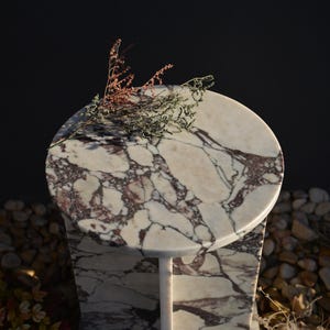May include: A small, round marble table with a white and burgundy veined pattern. The table has a single, slender leg and a decorative floral arrangement on top. The background is dark.