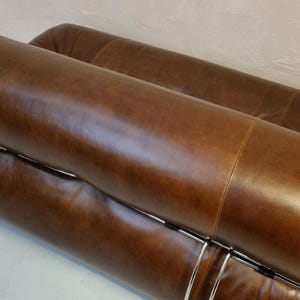 May include: Close-up of a brown leather sofa with a curved design. The sofa has a polished, smooth texture and is supported by a silver metal frame. The design is modern and minimalist, suitable for a living room.