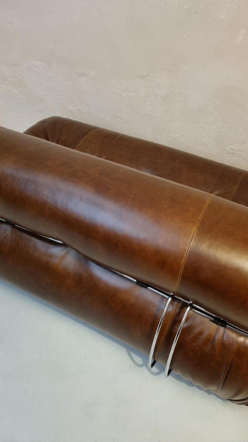 May include: Close-up of a brown leather sofa with a curved design. The sofa has a polished, smooth texture and is supported by a silver metal frame. The design is modern and minimalist, suitable for a living room.