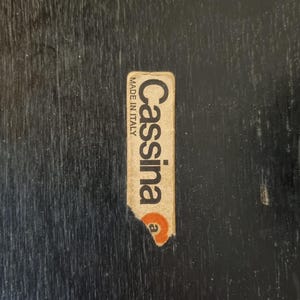 May include: A close-up of a black wooden surface with a Cassina Made in Italy sticker. The sticker is rectangular with the brand name in a serif font and a small orange circle.