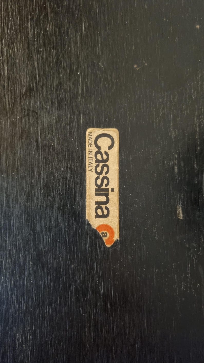 May include: A close-up of a black wooden surface with a Cassina Made in Italy sticker. The sticker is rectangular with the brand name in a serif font and a small orange circle.
