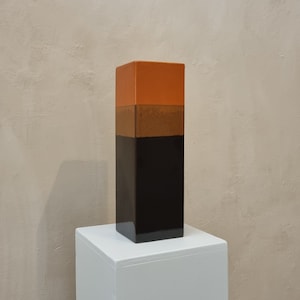 Op de afbeelding: Een rechthoekige, driekleurige decoratieve vaas. Het bovenste gedeelte is oranje, het midden is bruin en de onderkant is zwart. De vaas staat op een witte sokkel tegen een neutraal getinte muur.