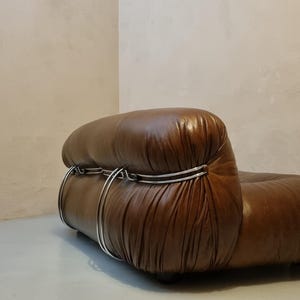 May include: A brown leather armchair with a rounded back and a low profile. The chair features a series of silver metal hoops that wrap around the back. The leather has a glossy finish. The chair is set against a neutral-toned wall.