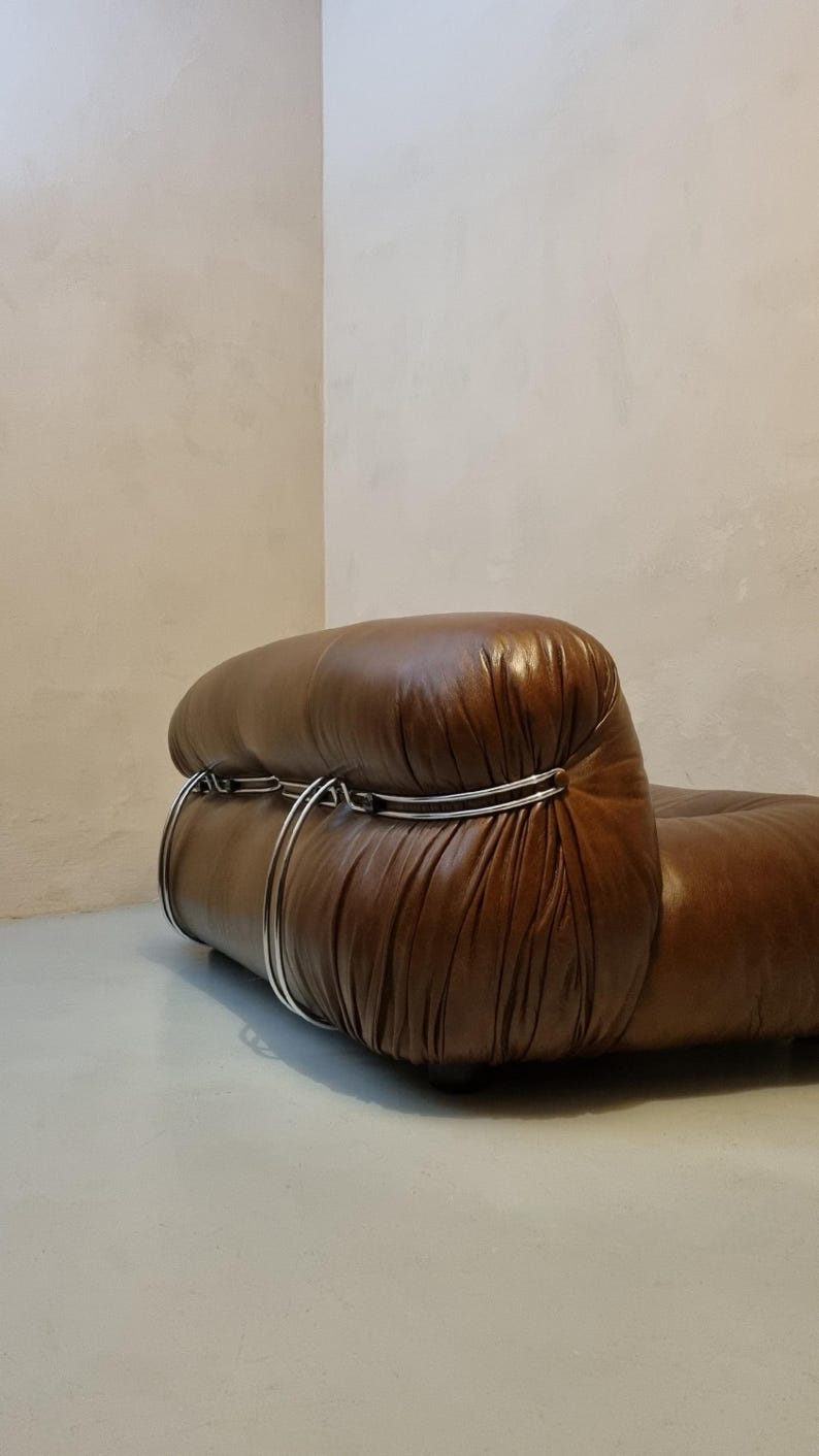 May include: A brown leather armchair with a rounded back and a low profile. The chair features a series of silver metal hoops that wrap around the back. The leather has a glossy finish. The chair is set against a neutral-toned wall.