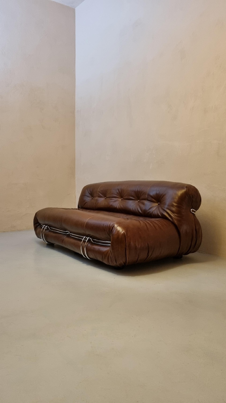May include: A brown leather sofa with a tufted back and rounded arms. The sofa has a low profile and is supported by a silver metal frame. The design is modern and minimalist, suitable for a living room or lounge area.