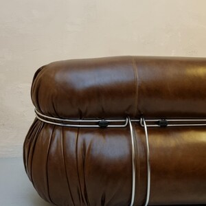 May include: A brown leather sofa with a rounded shape and a metal frame. The sofa has a tufted design and a dark brown color. The metal frame is silver and runs along the sides of the sofa.
