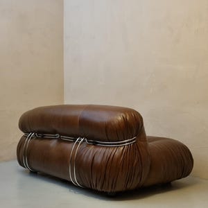 May include: A brown leather sofa with a rounded, low profile. The sofa features a tufted design and is accented with silver metal bands. The piece is set against a neutral-toned wall and floor, creating a minimalist aesthetic.