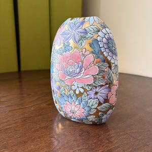 May include: An oval-shaped ceramic vase featuring a dense, colourful floral pattern. The design includes pink, blue, and purple flowers with green leaves set against a metallic gold background. The vase has a smooth, rounded silhouette and a flat top opening.
