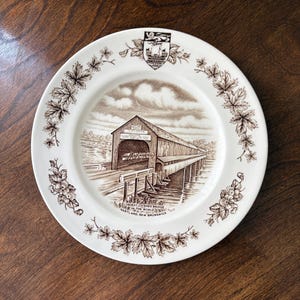 Könnte beinhalten: Ein weißer Keramikteller mit einer braunen Transferdruck-Illustration der Hartland Covered Bridge in New Brunswick. Der Rand ist mit einem braunen Ahornblattmuster und einem Wappen verziert. Der Text lautet: The Longest Covered Bridge in the World, Hartland, New Brunswick.