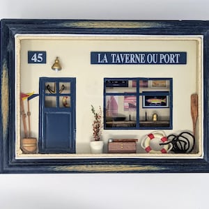 Puede incluir: Un marco de madera azul con un paspartú blanco y una escena en miniatura de una taberna francesa con un letrero que dice "La Taverne Ou Port". La escena incluye una puerta azul con el número "45" encima, una ventana con vista al mar, un salvavidas y una cuerda.