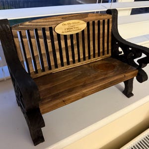 May include: A small wooden bench with a dark brown finish and black detailing. The back of the bench features vertical slats, and an oval plaque with the text "On My Silent Days I Miss You A Little Louder".