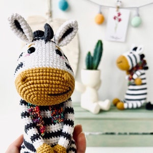 May include: Crocheted zebra figurine with black and white stripes, a mustard-colored muzzle, and a colorful scarf. The handmade stuffed animal has large ears and is approximately 6 inches tall. A second zebra figurine is in the background.