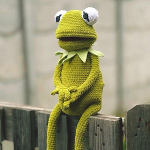 May include: A handmade, green crochet frog doll with white and black eyes, sitting on a wooden fence. The frog has long legs and arms, and a green collar. The frog is a whimsical, handcrafted item.