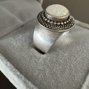 May include: A silver ring with a large, oval, iridescent opal stone. The stone is set in a detailed bezel with a textured band. The ring is displayed in a gray velvet ring box.