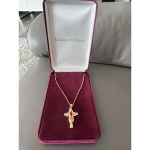 May include: A gold-tone cross pendant necklace featuring a central marquise-cut red stone and small clear accent stones, presented in a burgundy velvet-lined display box with the text Camrose & Kross printed on the inside lid of the case.