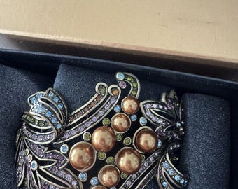 Puede incluir: Un brazalete de puño ornamentado con un diseño floral. El brazalete presenta metal de color bronce, adornado con brillantes pedrería en tonos de azul, morado, verde y dorado. El brazalete se presenta en una caja con el texto "HEIDI DAUS".