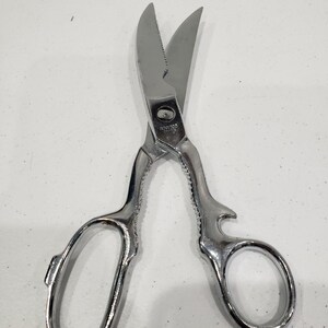 May include: Stainless steel kitchen shears with curved blades and ergonomic handles. The shears have a serrated edge on one blade and a smooth edge on the other. The handles have finger loops for a secure grip.