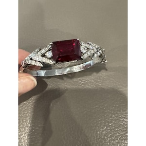 May include: A silver-toned hinged bangle bracelet featuring a large, rectangular, deep red center stone. The band is embellished with small, clear, sparkling stones arranged in a leaf-like pattern on both sides of the central red gemstone.