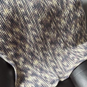 May include: A handmade crochet blanket in shades of blue, cream, and gray. The blanket features a textured pattern with diagonal stripes and decorative stitch details. The soft, cozy blanket is draped over a black surface.