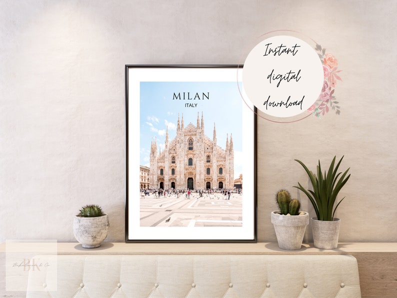 May include: A printable digital download of a photograph of the Duomo di Milano, a large cathedral in Milan, Italy. The image is in a black frame and is displayed on a shelf with plants.
