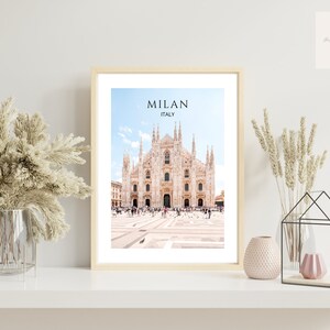 May include: A framed print of the Duomo di Milano, a large cathedral in Milan, Italy. The print features a photo of the cathedral with a blue sky and white clouds in the background. The text "MILAN ITALY" is printed at the top of the print.