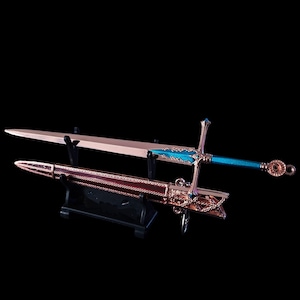 Puede incluir: Una espada decorativa con una empuñadura dorada y una hoja azul, expuesta en un soporte negro. La espada tiene detalles intrincados en la empuñadura y una vaina.