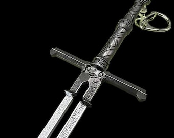 May include: A silver-coloured sword keychain with intricate details and a black handle. The sword has a double-edged blade and a decorative crossguard.