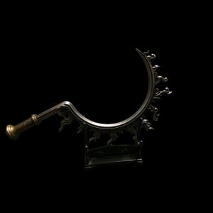 May include: A dark metallic sickle with a curved blade featuring ornate, flame-like decorative protrusions along the outer edge. The handle is cylindrical with a flared end, and the entire object is supported by a rectangular stand.