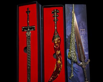May include: A set of three decorative swords displayed in individual boxes. The first is a dark metallic sword with intricate filigree, the second is a curved blade with red and gold patterns, and the third is a textured silver and bronze sword.