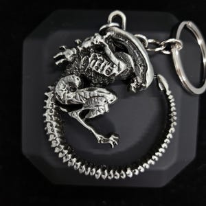 May include: A silver-colored metal keychain featuring a detailed alien creature with a curled tail. The alien is in a curled position with its head and body facing forward. The keychain is attached to a silver-colored metal ring.