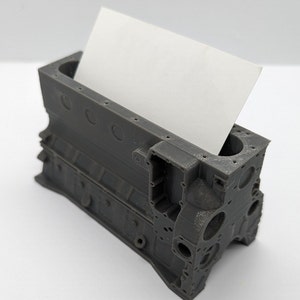 Puede incluir: Un modelo de bloque de motor impreso en 3D de color gris, diseñado para guardar tarjetas de visita. El modelo tiene un diseño realista con detalles intrincados y una ranura para insertar tarjetas.