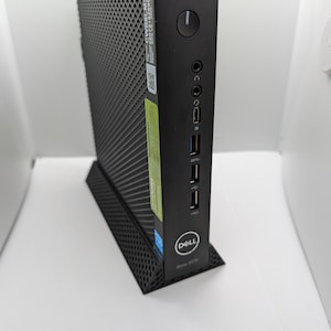 May include: A black Dell Wyse 5070 thin client with a black stand. The client has a power button, a USB port, a headphone jack, and a display port. The client has a black and gray textured surface.