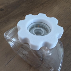 May include: A clear, graduated container with a white, gear-shaped lid. The lid sits atop a clear, transparent container with measurement markings. The container is on a wooden surface.