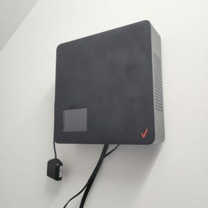 May include: A black rectangular device with a small screen and the Verizon logo, mounted on a white wall. The device has a perforated side and is connected to a power adapter and cables.