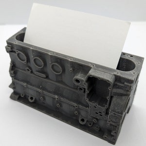 Puede incluir: Un modelo de bloque de motor impreso en 3D de color gris, diseñado para contener tarjetas de visita. El modelo tiene un diseño realista con detalles intrincados y una superficie texturizada.