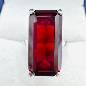 May include: A rectangular-cut, deep red gemstone ring with a silver band. The gemstone is set with four silver prongs. The ring is displayed in a white jewelry box with a blue interior.