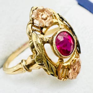 May include: A gold ring featuring a central oval-cut vibrant red gemstone set in a bezel. The band and setting are decorated with textured gold floral motifs and leaf-like engravings, creating an intricate, multi-toned metal design.