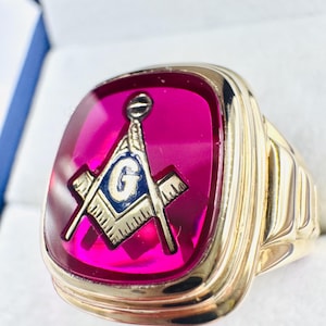 May include: Gold-tone ring with a large, rectangular, magenta-colored stone. The stone features a Masonic symbol with a compass, square, and the letter "G". The ring has a detailed, ridged band.