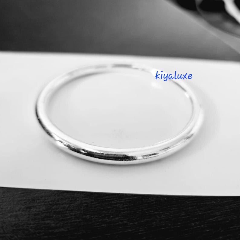 May include: A shiny, silver ring with a smooth, rounded design. The ring is set against a white background, with the word "kiyaluxe" in blue script. The ring's simple, elegant style suggests it could be a wedding band or a fashion accessory.