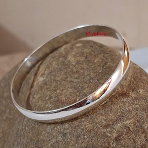 May include: A shiny, silver bangle bracelet with a smooth, rounded design. The bracelet reflects the surrounding environment, creating a mirrored effect. The word "Kiyaluxe" is visible on the bracelet.