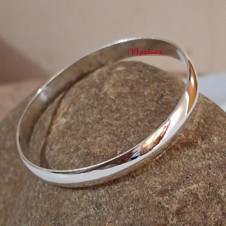 May include: A shiny, silver bangle bracelet with a smooth, rounded design. The bracelet reflects the surrounding environment, creating a mirrored effect. The word "Kiyaluxe" is visible on the bracelet.
